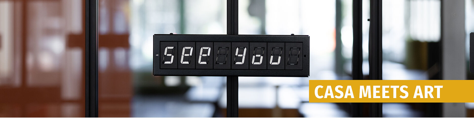 Electronic display board showing the text 'see you' against a blurred background with glass and wood elements.