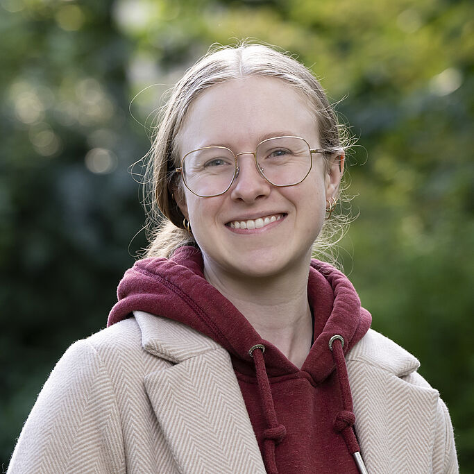 Portrait of a young person with light hair wearing a light jacket and a dark red hoodie against a blurred green background. It is Erica blum.