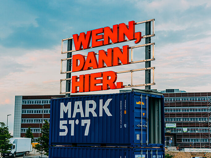 A neon sign with the words “If anywhere, then here” and “MARK 57°” on a building in an urban setting
