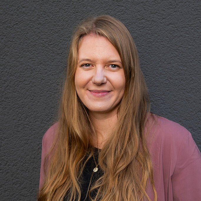 Woman with long loose blonde hair smiling in front of a dark gray wall wearing a black blouse and a pink cardigan