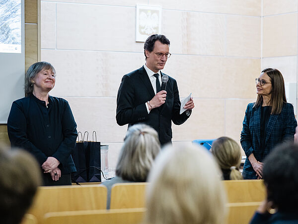 Drei Personen stehen vor einem Publikum in einem Vorleseungssaal. In der Mitte steht NRW Ministerpräsident Hendrik Wüst, links daneben CASA PI Angela Sasse.