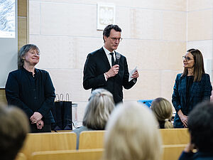 Drei Personen stehen vor einem Publikum in einem Vorleseungssaal. In der Mitte steht NRW Ministerpräsident Hendrik Wüst, links daneben CASA PI Angela Sasse.