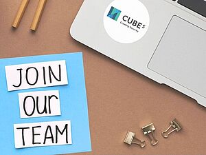Laptop mit Aufkleber 'CUBE 5 Creating Security', daneben Zettel mit handgeschriebenem Text 'JOIN OUR TEAM', zwei Bleistifte und drei Büroklammern auf braunem Untergrund
