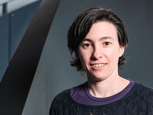 Porträt einer lächelnden Frau mit kurzen dunklen Haaren vor einem dunklen Hintergrund mit diagonalen Linien