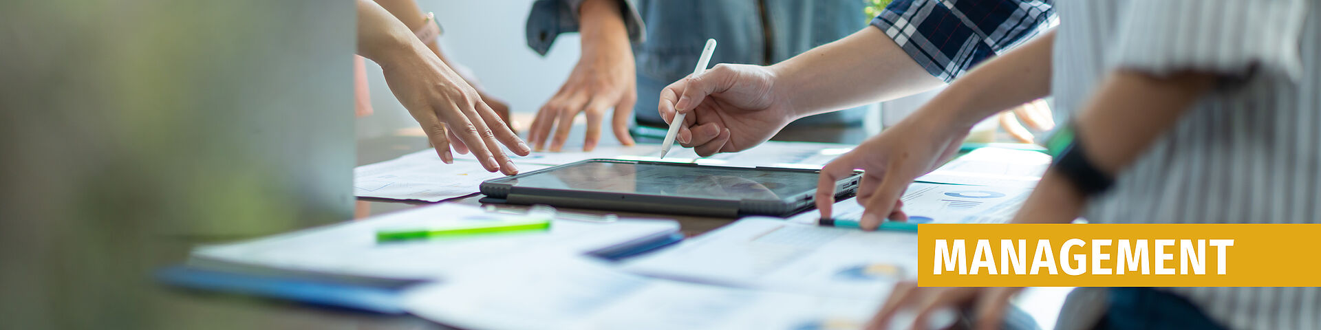 Mehrere Hände über einem Tisch mit Tablet und Papieren, eine Hand hält einen Stift, rechts oben steht in gelbem Kasten 'MANAGEMENT'
