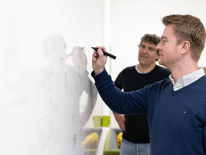 Two men stand in front of a white wall, one writing on it with a marker