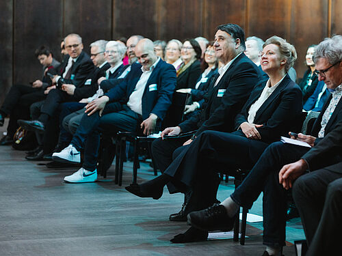Das Publikum auf Stuehlen darunter Rektor Martin Paul und Ministerin Ina Brandes