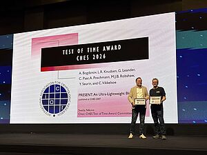 Two men standing on a stage holding certificates in front of a large screen displaying the 'Test of Time Award CHES 2026' with names and a logo
