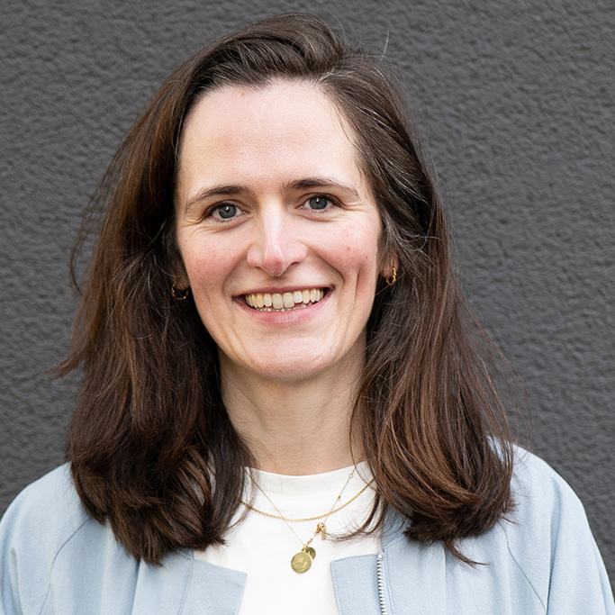 Frau mit schulterlangen braunen Haaren, weißem Hemd und goldener Halskette vor grauem Hintergrund. Es handelt sich um Johanna Luttermann.